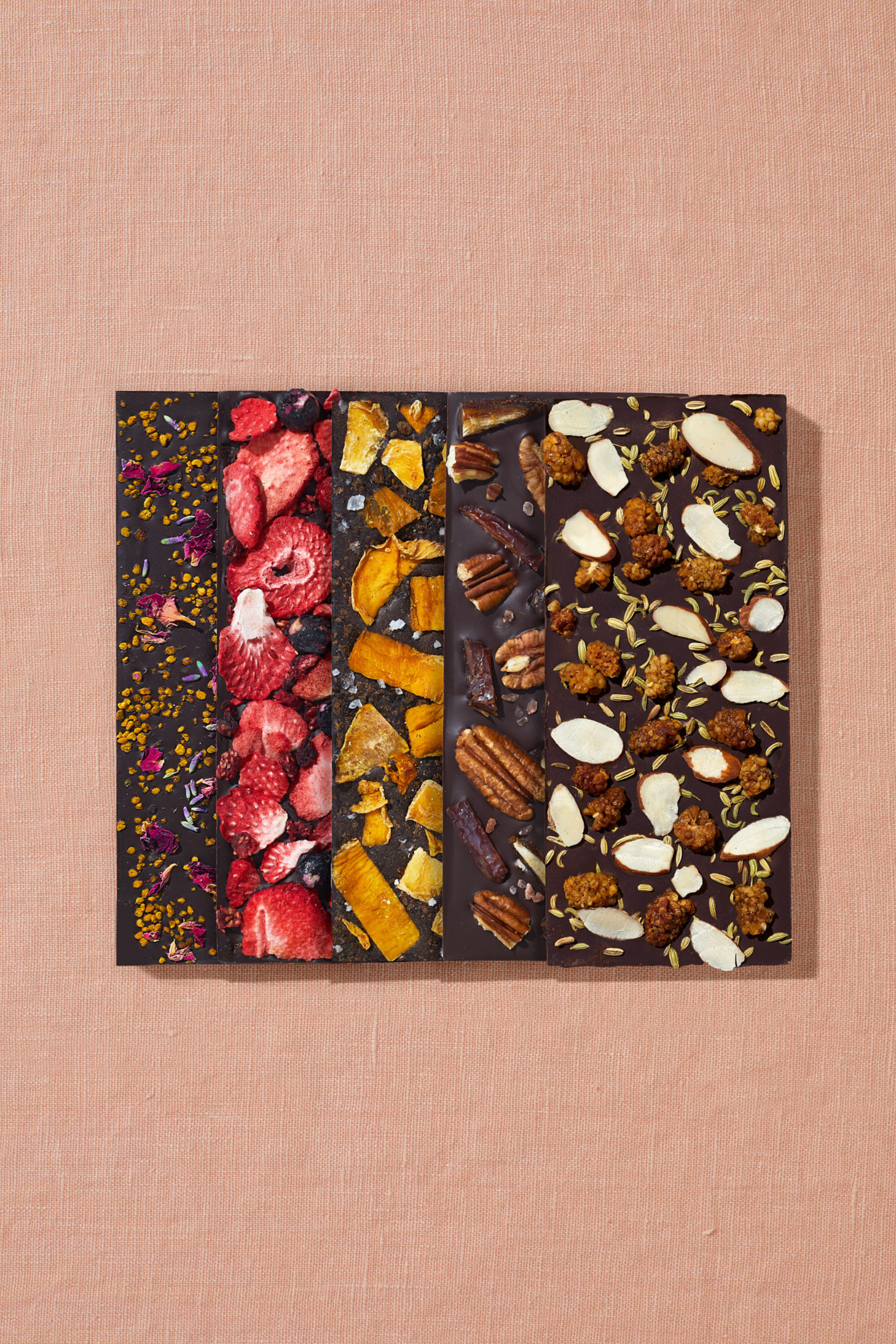 A set of 5 date sweetened chocolate bars with no added sugar is arranged in a flay lay upon peach linen fabric. Warm, direct light hits the bars, which go from left to right, starting with Lavender, Bee Pollen, Rose Petal; Mixed Berry; Medjool Date, Pecan, Himalayan Salt; Mango, Urfa Chili, Black Lime; and, White Mulberry, Almond, Fennel.