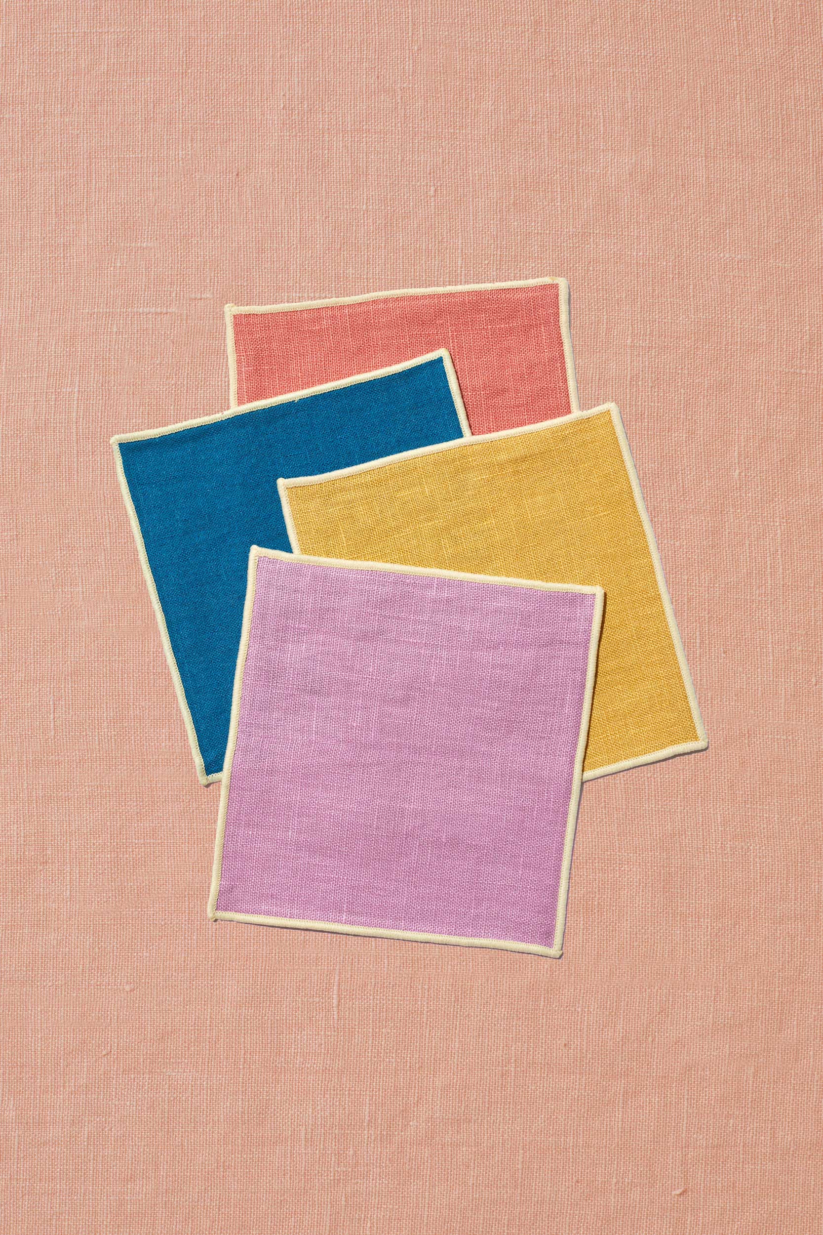 A set of four dessert napkins sits upon a peach linen background. The napkins are artfully scattered with rose in the back, followed by marine blue, mustard, and lavender in front. Each napkin matches the colors of Spring & Mulberry's date sweetened chocolate boxes, which are inspired by the setting sun.
