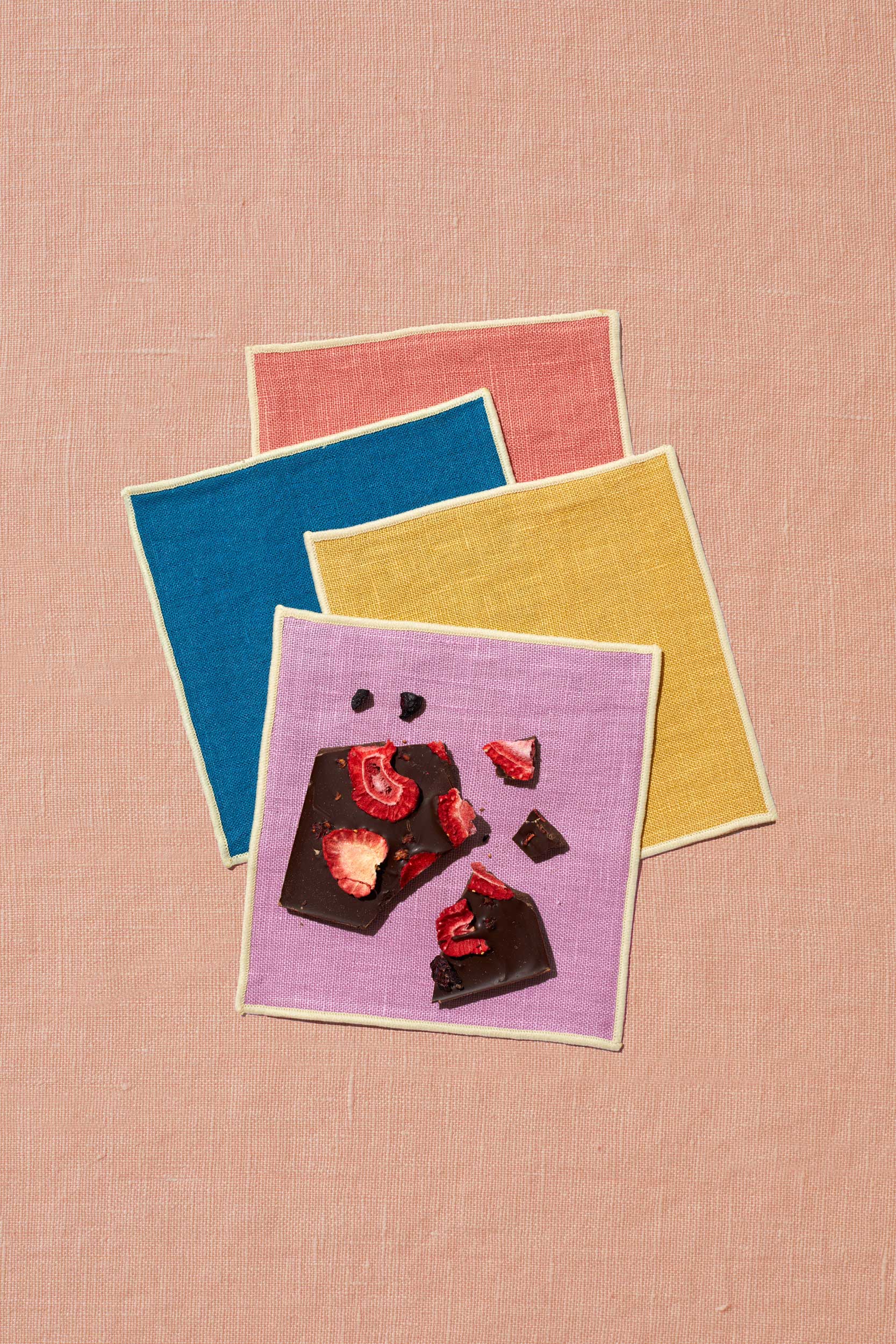A set of four napkins sits upon a peach linen background. The napkins are artfully scattered and come in four colors with ivory trim: rose, marine blue, mustard, and lavender. On the front napkin, which is lavender, sits a broken bar of date sweetened chocolate topped with mixed berries. The chocolate has no added sugar.