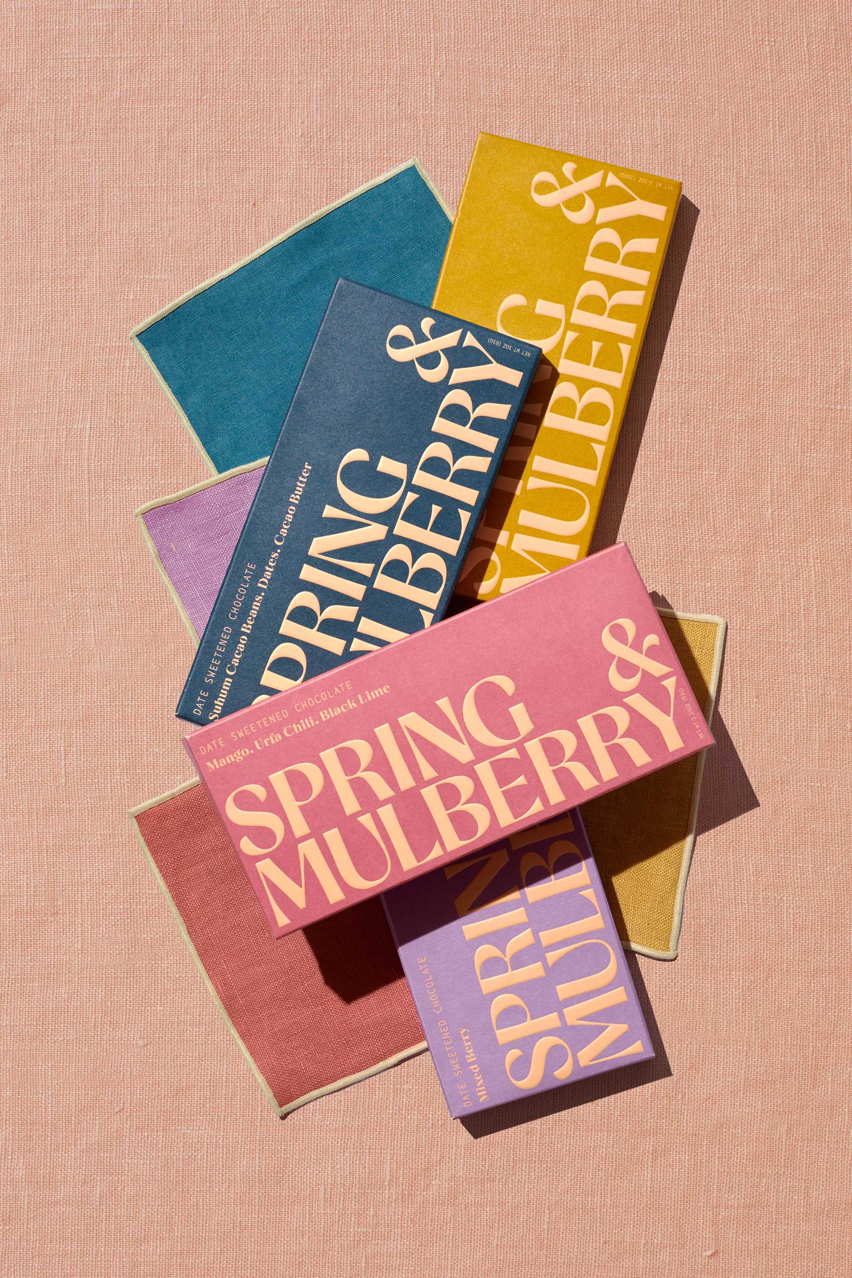 Four date sweetened chocolate bar boxes in rose, marine blue, mustard, and lavender sit artfully displayed upon four 5" dessert napkins in corresponding hues. The boxes contain no sugar added chocolate in Mango, Urfa Chili, Black Lime; Pure Dark; Medjool Date, Pecan, Himalayan Salt; and, Mixed Berry. The napkins and chocolate bars are sold as a set.