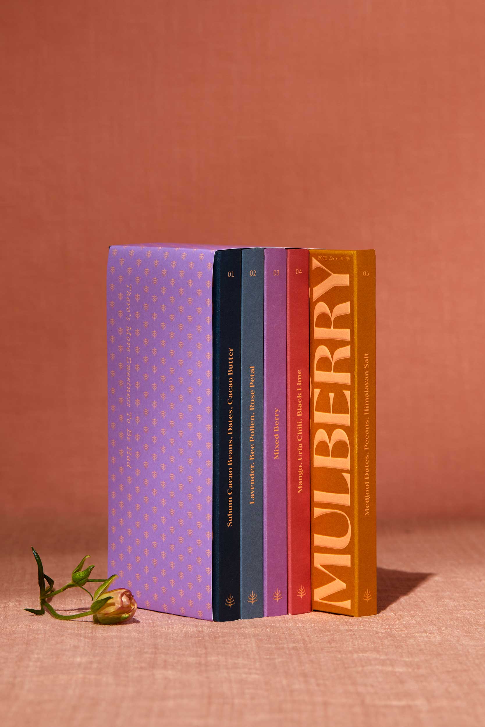 A gift set of 5 date sweetened chocolate bars with no added sugar stands on a peach linen backdrop. To the left of the gift set is a delicate flower. The gift set resembles a library set of books and is wrapped in purple paper adorned with a mulberry motif. The 5 chocolate boxes include: Pure Dark in a navy box; Lavender, Bee Pollen, Rose Petal in a sky blue box; Mixed Berry in a purple box; Mango, Urfa Chili, Black Lime in a rose box; and, Medjool Date, Pecan, Himalayan Salt in a mustard box.