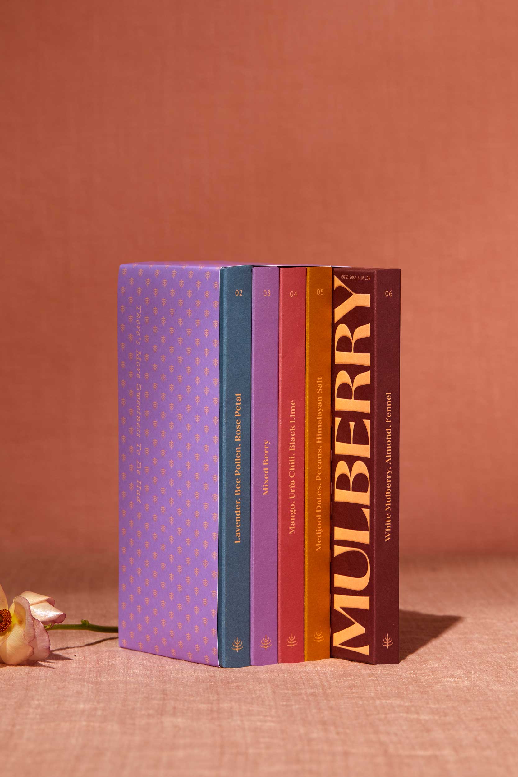 A gift set of 5 date sweetened chocolate bars with no added sugar stands on a peach linen backdrop. To the left of the gift set is a delicate flower. The gift set resembles a library set of books and is wrapped in purple paper adorned with a mulberry motif. The 5 chocolate boxes include: Lavender, Bee Pollen, Rose Petal in sky blue; Mixed Berry in lavender; Mango, Urfa Chili, Black Lime in rose; Medjool Date, Pecan, Himalayan Salt in mustard; and, White Mulberry, Almond, Fennel in a maroon box.