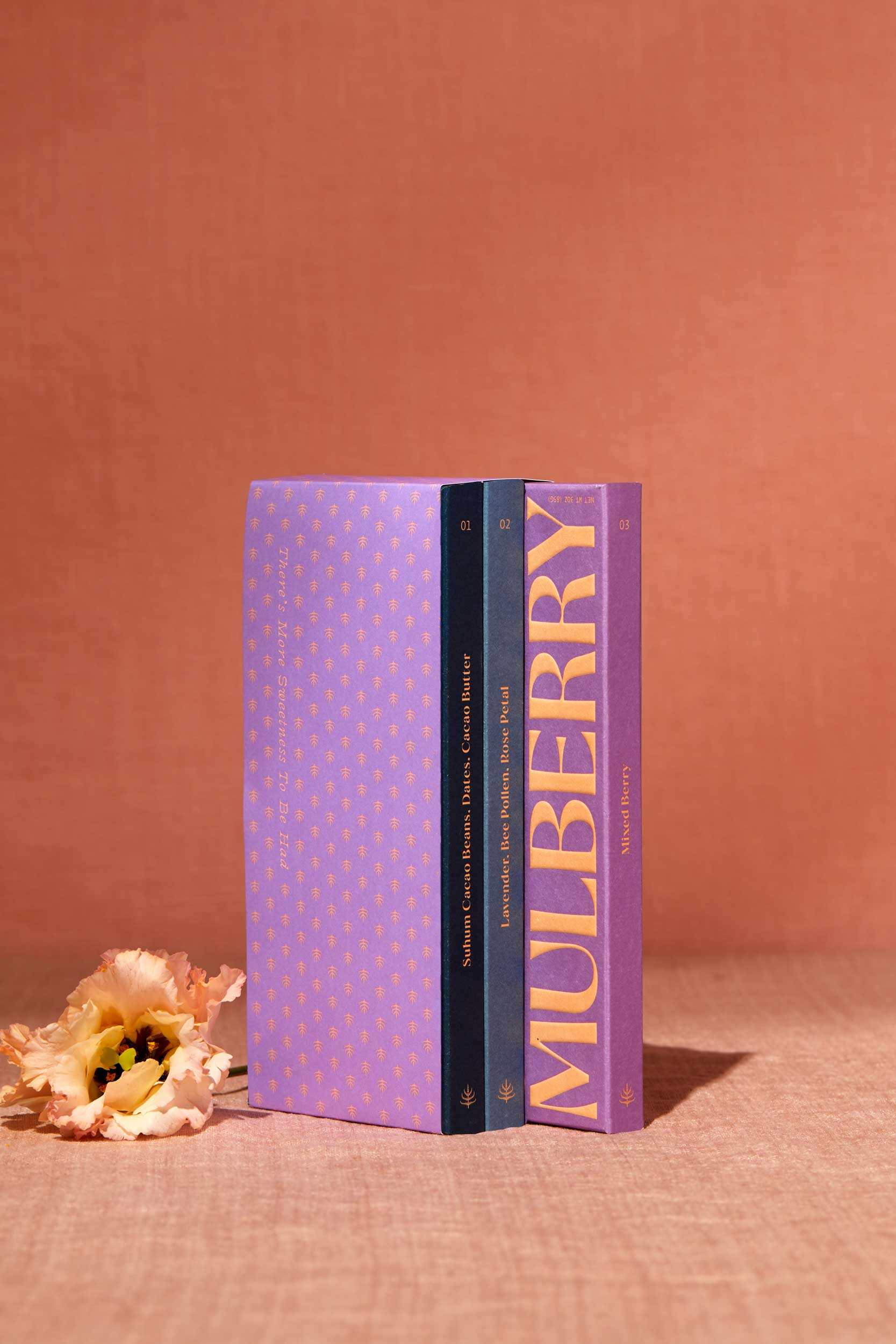 A gift set of 3 date sweetened chocolate bars with no added sugar stands on a peach linen backdrop. To the left of the gift set is a delicate flower. The gift set resembles a library set of books and is wrapped in purple paper adorned with a mulberry motif. The 3 chocolate boxes include: Pure Dark in a navy box; Lavender, Bee Pollen, Rose Petal in a sky blue box; and, Mixed Berry in a purple box.