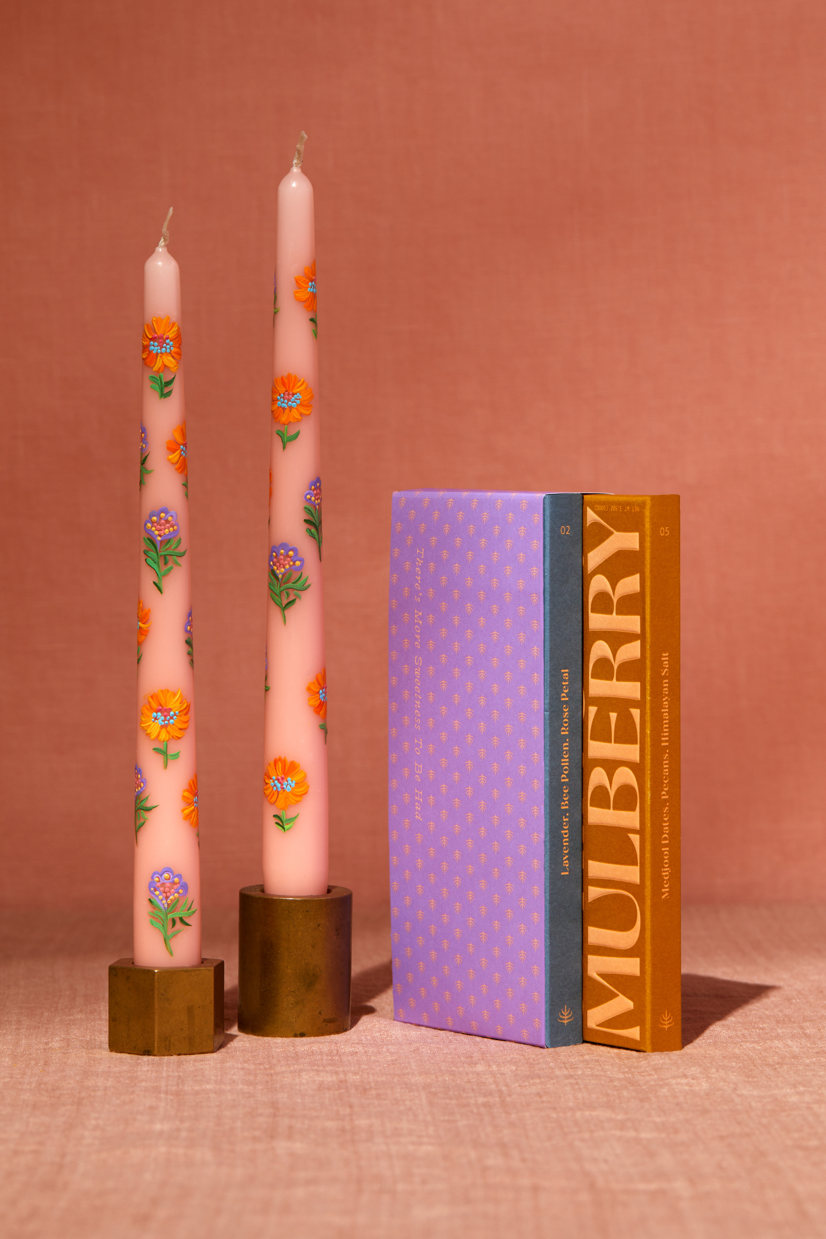A set of two 10" tapered peach candles stands in brass candlesticks. Each candle is adored with handpainted flowers in peach and lavender shades. To the right is a gift set of 2 chocolate bars wrapped in a mulberry printed lavender gift paper. The bars contain Lavender, Rose Petal, Bee Pollen date sweetened chocolate and Medjool Date, Pecan, Himalayan Salt date sweetened chocolate, both with no added sugar. The image is shot straight on with warm light on a peach backdrop.